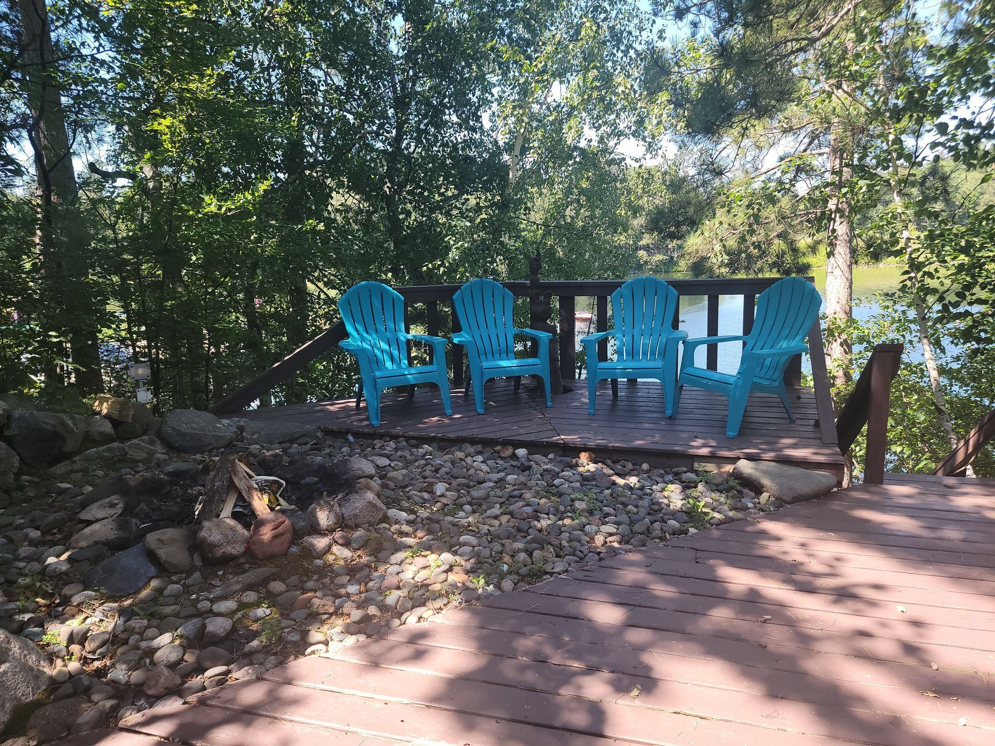 A wooden deck with four blue Adirondack chairs, a stone wall, and a view of a lake.