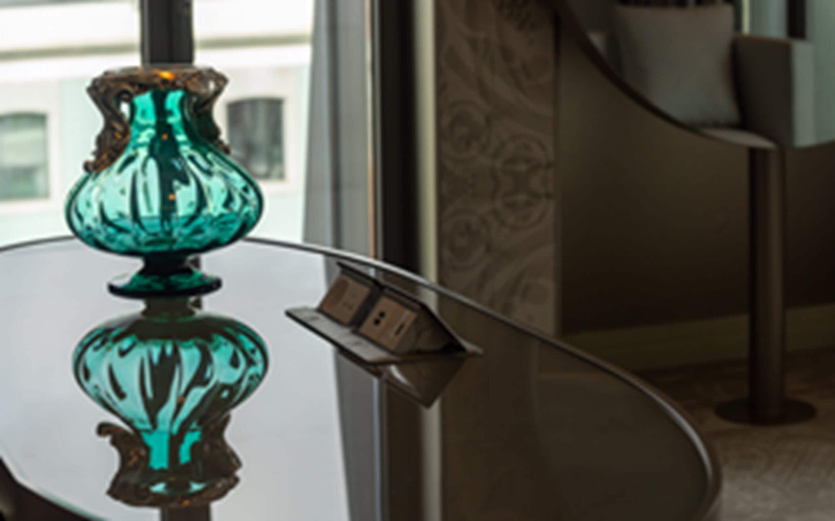 A teal glass vase on a reflective glass table.