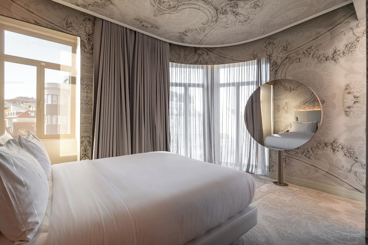 A bedroom with a large bed, a round mirror, and a view of the city through large windows.