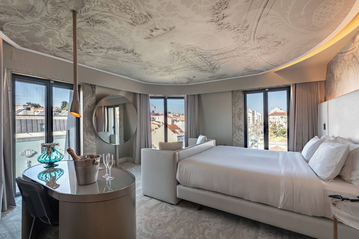 A modern hotel room with a large bed, a seating area, a dining table, and a view of the cityscape.