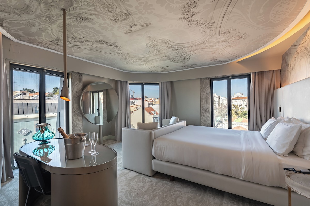 Una habitación de hotel moderna con una cama grande, un área de descanso, una mesa de comedor y vistas a la ciudad.