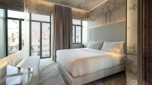 Habitación de hotel con una cama grande, zona de estar con una mesa de vidrio y un balcón con vista a edificios.
