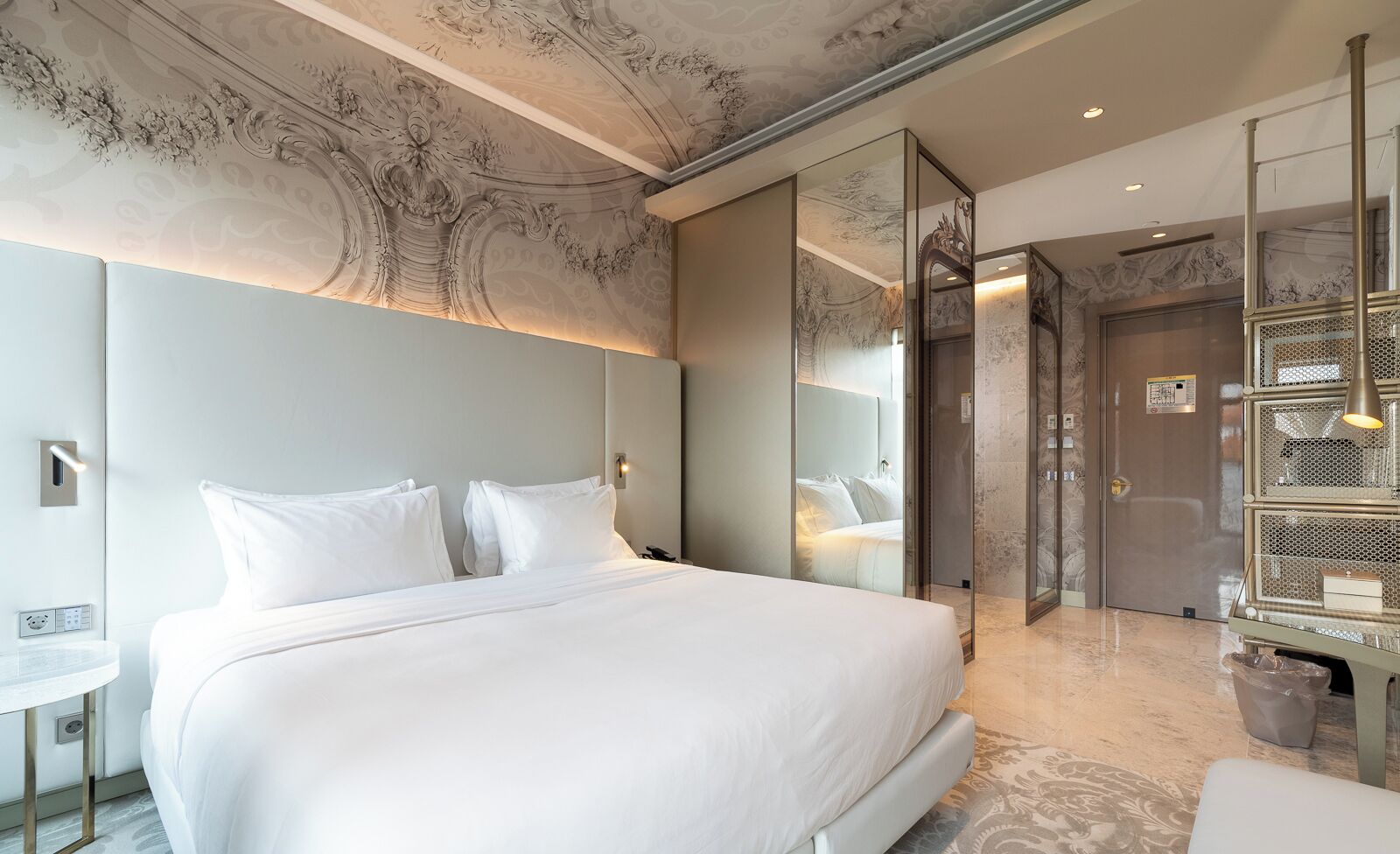 A modern hotel room with a large bed, a bedside table, a mirror, and a glass-enclosed elevator.