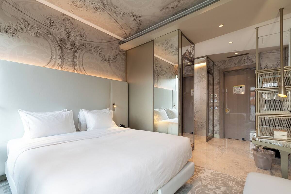 A modern hotel room with a large bed, a bedside table, a mirror, and a glass-enclosed elevator.