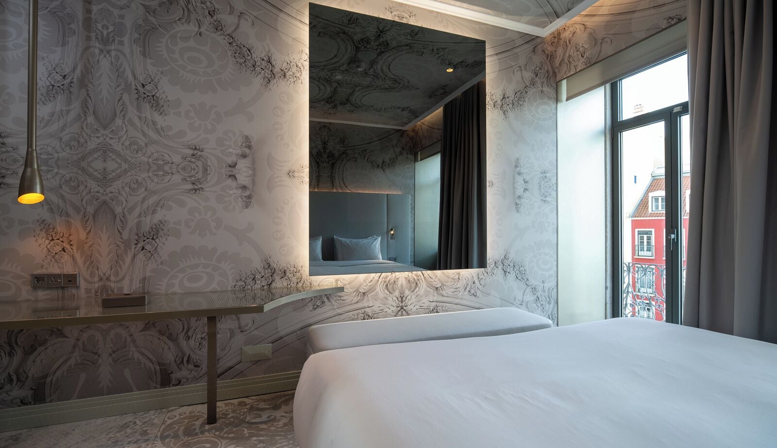 A hotel room with a bed, a bedside table, a mirror, and a window with a view of buildings.