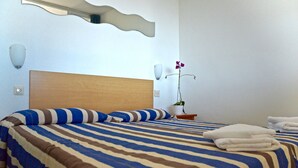 Quarto de hotel com uma cama grande, roupa de cama listrada, cabeceira de madeira, mesa de cabeceira com uma planta e luminária fixada na parede.