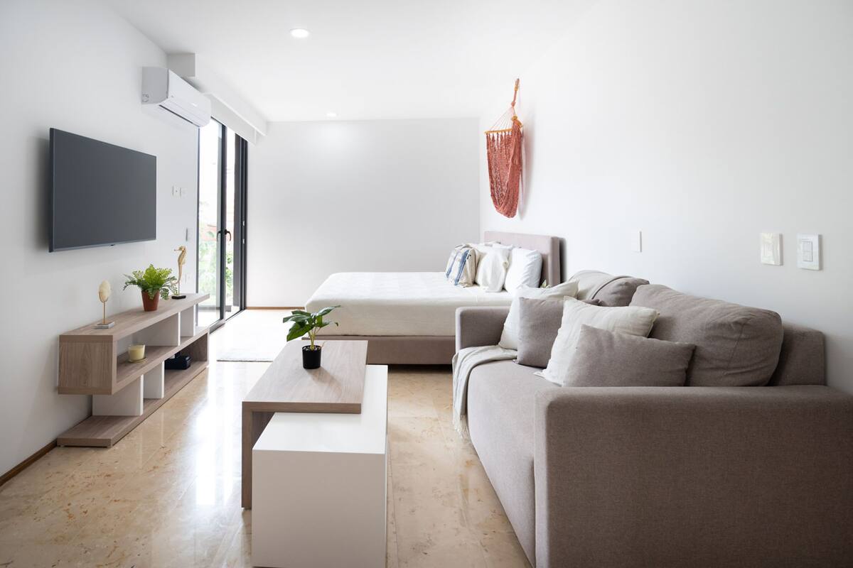 Un dormitorio moderno y minimalista con cama, sofá, televisor y una planta.