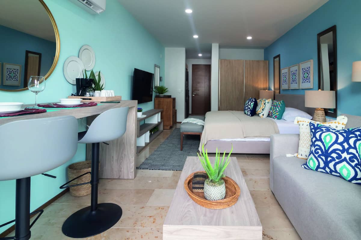Una sala moderna con sofá, mesa de centro y televisor. Hay una zona de comedor con sillas y una mesa. La habitación tiene una pared azul y un mueble de madera.