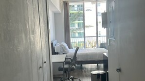A hotel room with a bed, a desk, a chair, a trash bin, and a view of a building through a window. - MIA Edgewater with sea view. (Miami)