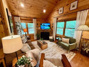 A cozy cabin interior with a fireplace, comfortable seating, and large windows offering a view of the outdoors. - Rustic Pines I One-Level Cabin Near Blue Ridge (Morganton)