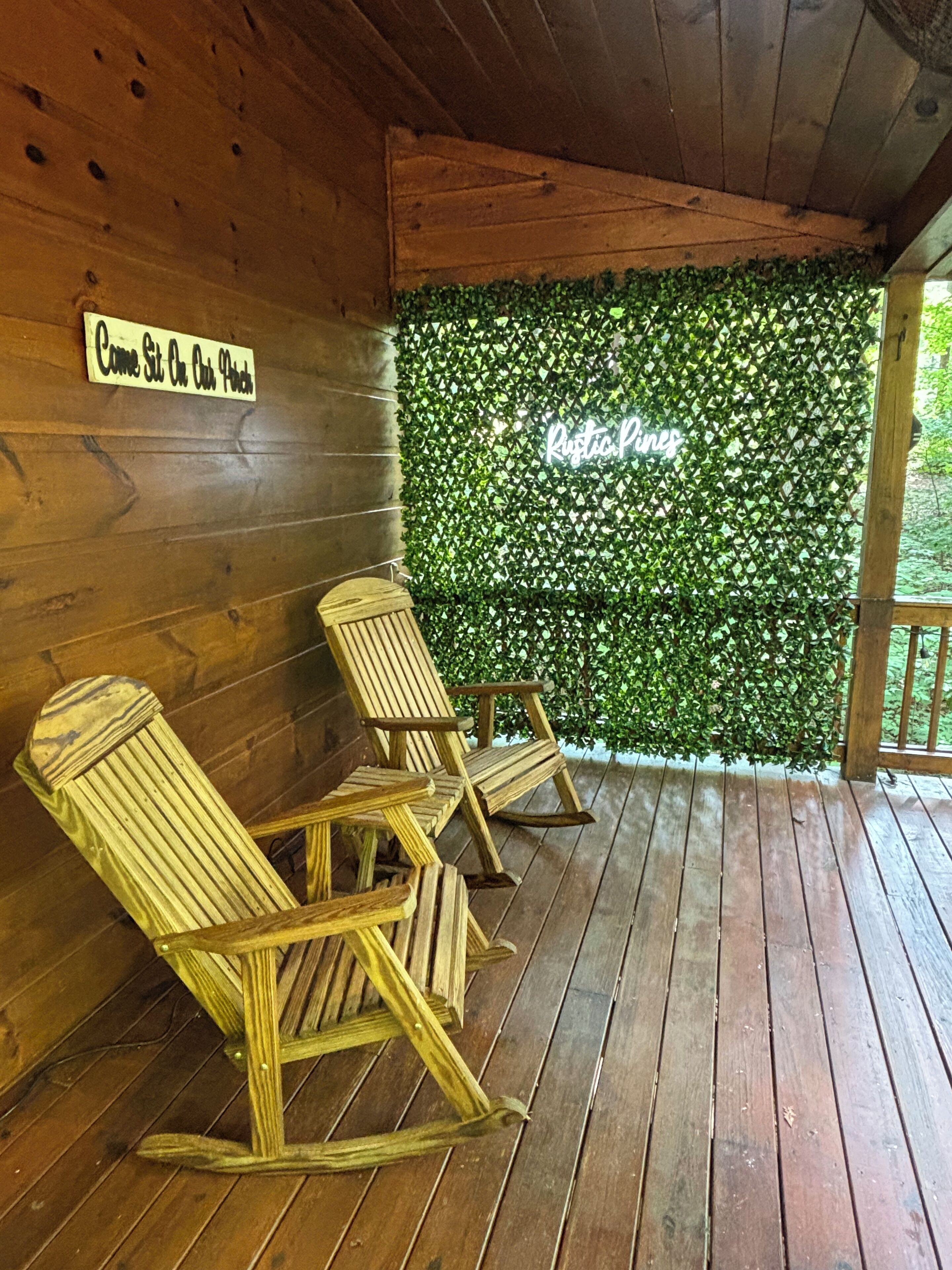 Une cabane en bois avec une véranda, deux fauteuils à bascule en bois, une enseigne accrochée au mur et un mur recouvert de verdure.