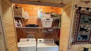 Une buanderie avec deux machines à laver et deux sèche-linge à chargement frontal, tous deux blancs. Des étagères en bois, situées au-dessus, servent à ranger les produits de nettoyage et un panier. Un ours en peluche décore le mur.