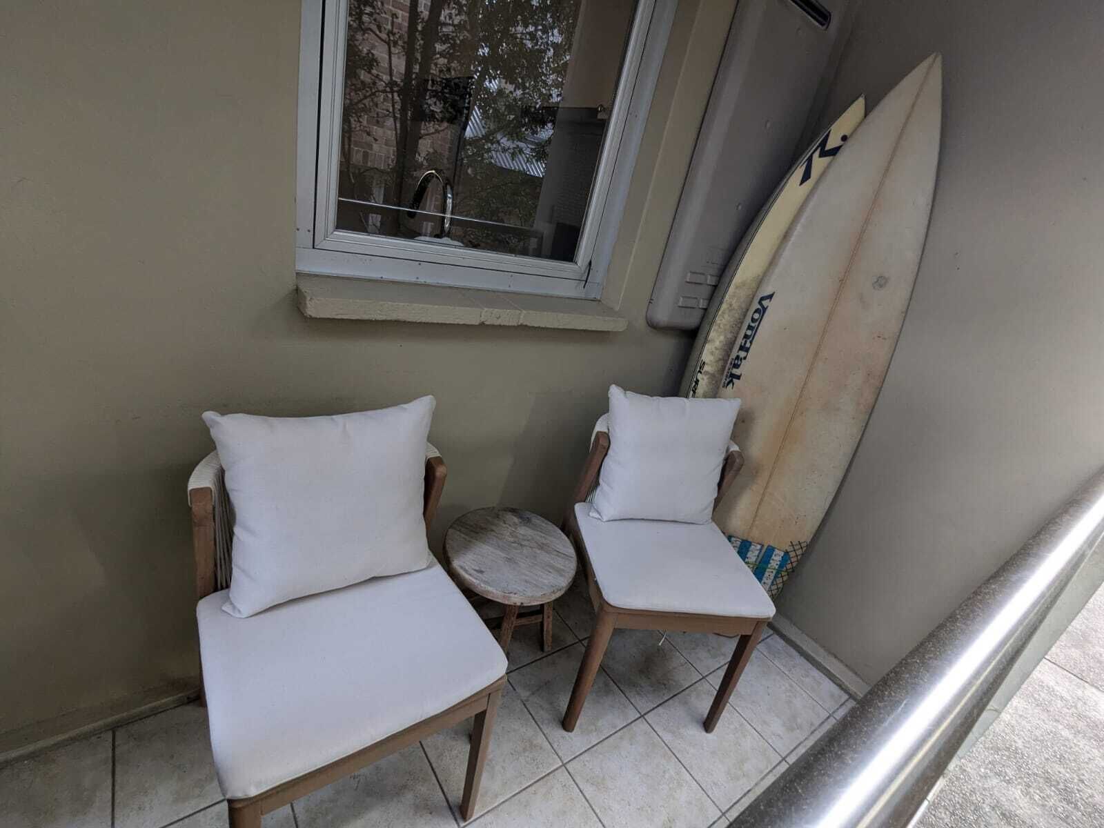 Un petit coin salon avec deux chaises en bois et une table ronde, une planche de surf appuyée contre le mur et une fenêtre donnant sur un immeuble.
