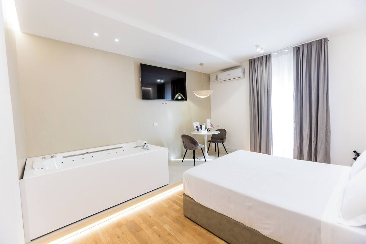 Habitación de hotel con una cama grande, bañera integrada, una pequeña mesa con sillas, televisor montado en la pared y un equipo de aire acondicionado.