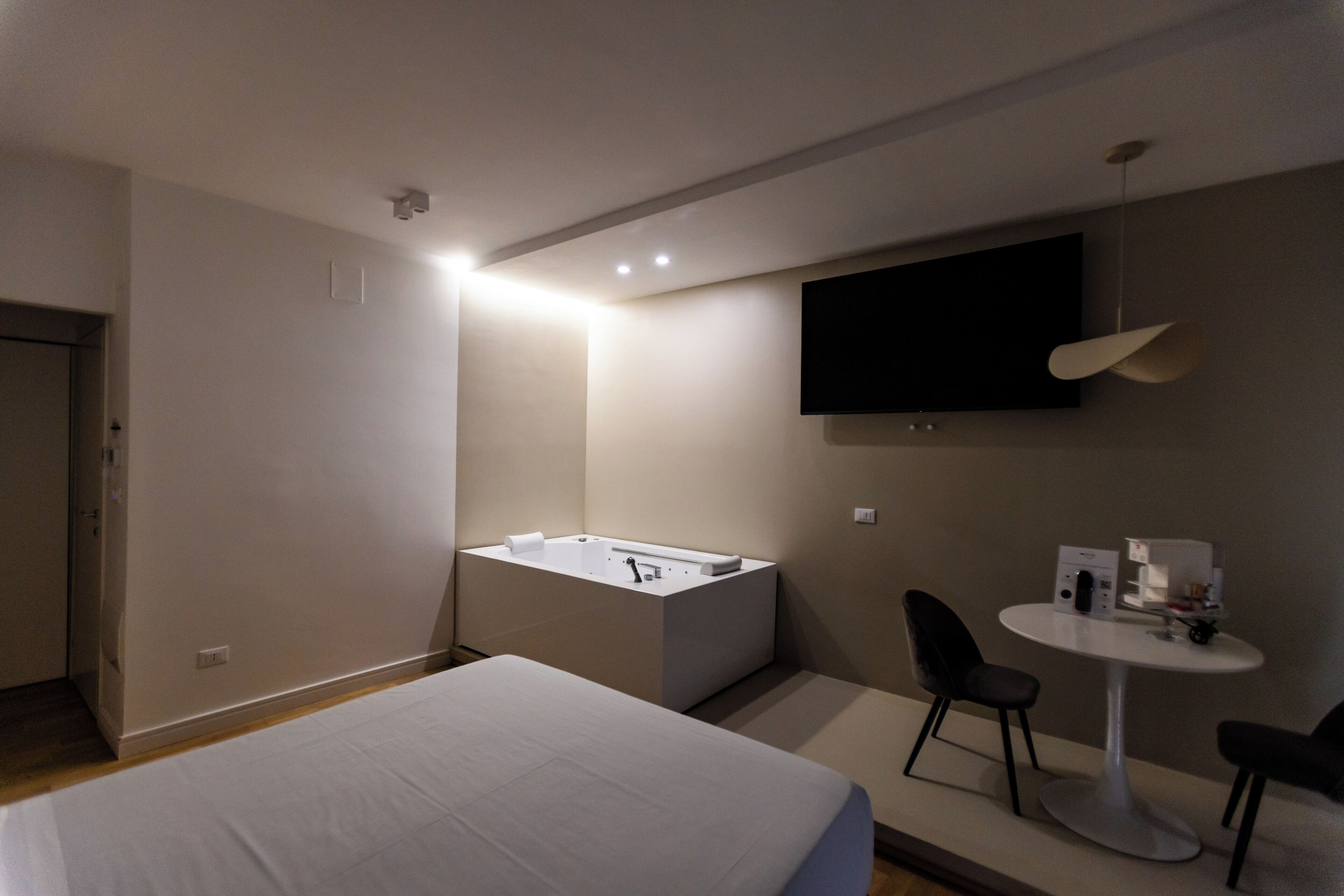 Una habitación de hotel moderna con una cama grande, una pequeña zona de estar con una mesa y sillas, una bañera empotrada y un televisor de pantalla plana montado en la pared.