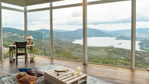 Een ruime kamer met grote ramen en een panoramisch uitzicht op een meer en omliggende heuvels.