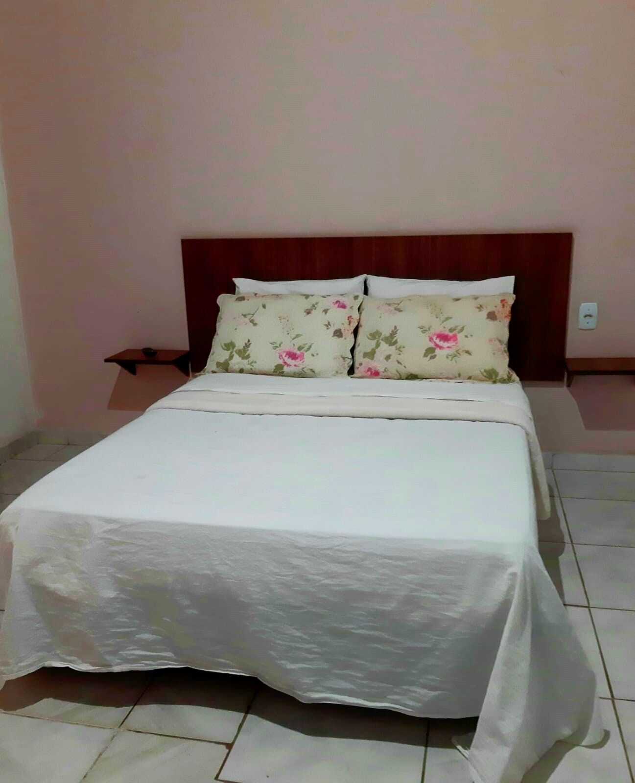 A neatly made bed with floral pillows and a white bedspread in a room with a tiled floor and a wooden headboard.