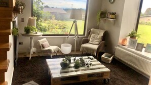 Une pièce chaleureuse avec une grande fenêtre, une table basse en bois, une chaise, une lampe et une petite étagère avec des plantes.