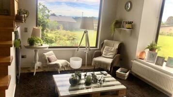 A cozy room with a large window, a wooden coffee table, a chair, a lamp, and a small shelf with plants.
