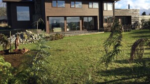 Une maison moderne à deux étages, dotée de grandes fenêtres, d’une cheminée en pierre et d’une pelouse bien entretenue.