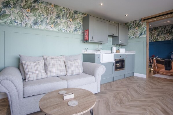 A modern living room with a grey sofa, a round coffee table, and a kitchen area with light blue cabinetry. - Brandy Cove - 1 Bed Cabin - Landimore (Swansea)