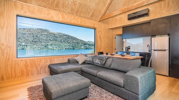 A modern cabin interior with a large window overlooking a lake and mountains, a gray sectional sofa, a small ottoman, and a kitchen area with stainless steel appliances. - Picture Perfect - Queenstown Holiday Home (Queenstown)