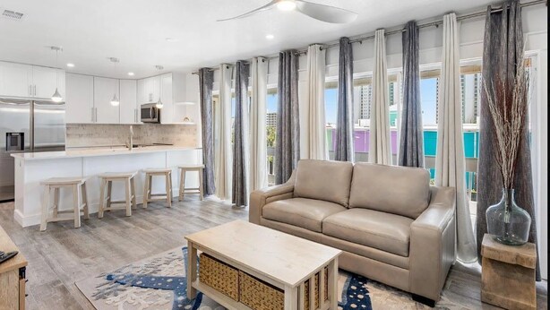 Une cuisine moderne avec des meubles blancs, un îlot central doté de tabourets, un canapé beige et un ventilateur de plafond.