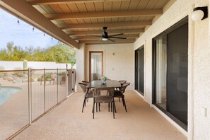 En overdækket terrasse med et bord og stole, en ventilator i loftet og udsigt til en swimmingpool.