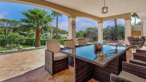 A covered outdoor dining area with wicker furniture and a glass-top table, surrounded by a garden with palm trees and a view of the landscape. - Villa Diamond (Quarteira)