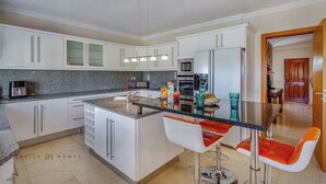 A modern kitchen with white cabinets, a granite countertop, and a central island with bar stools. - Villa Diamond (Quarteira)
