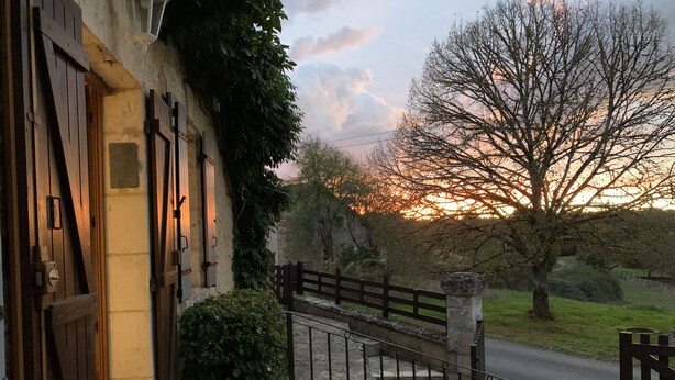 Ein Steinhaus mit einer Holztür und einem Metallgitter, einer Steintreppe und Blick auf einen Sonnenuntergang über einem Baum und einer Wiese.