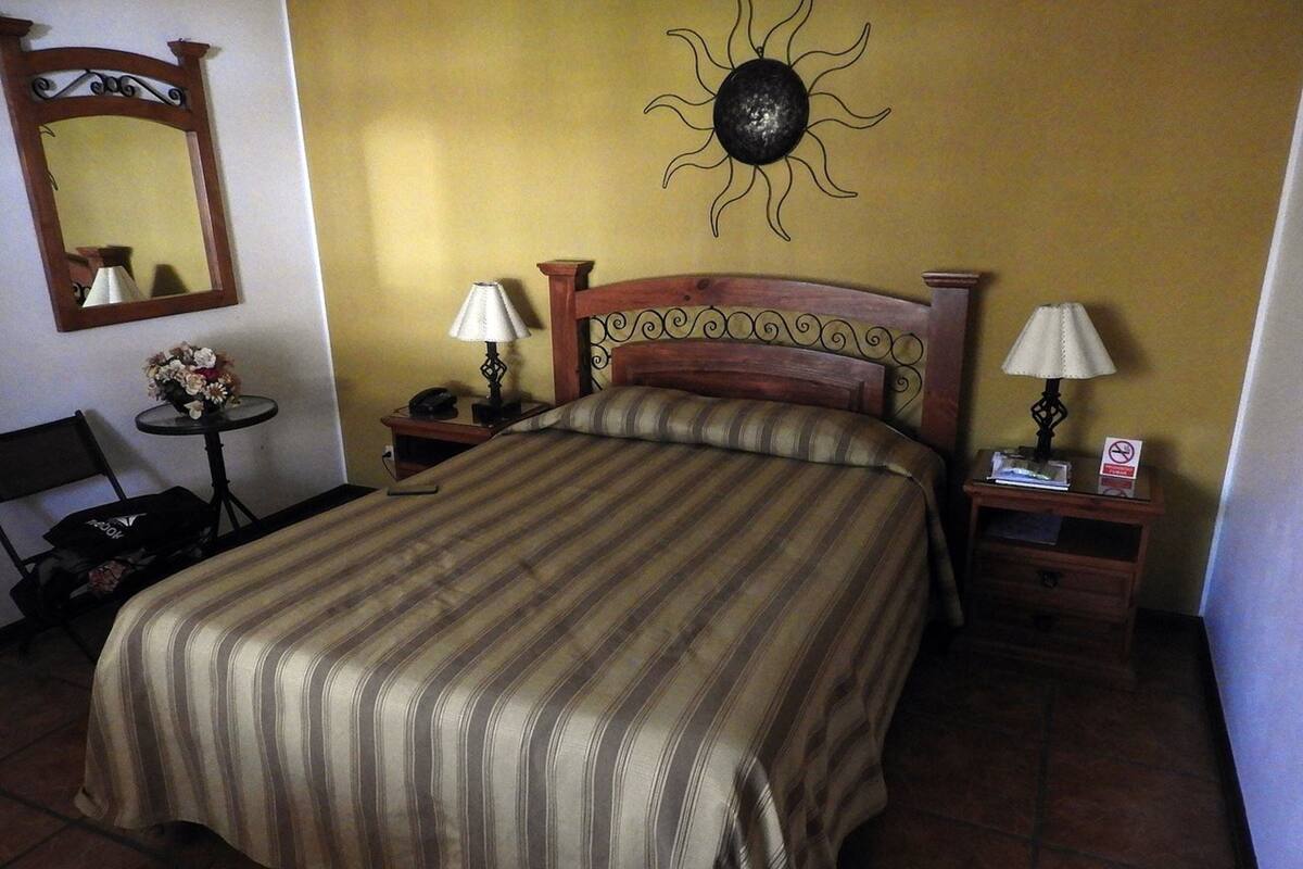 A hotel room with a large bed, two bedside tables with lamps, a small table with a vase of flowers, a chair, and a wall-mounted sun decoration.