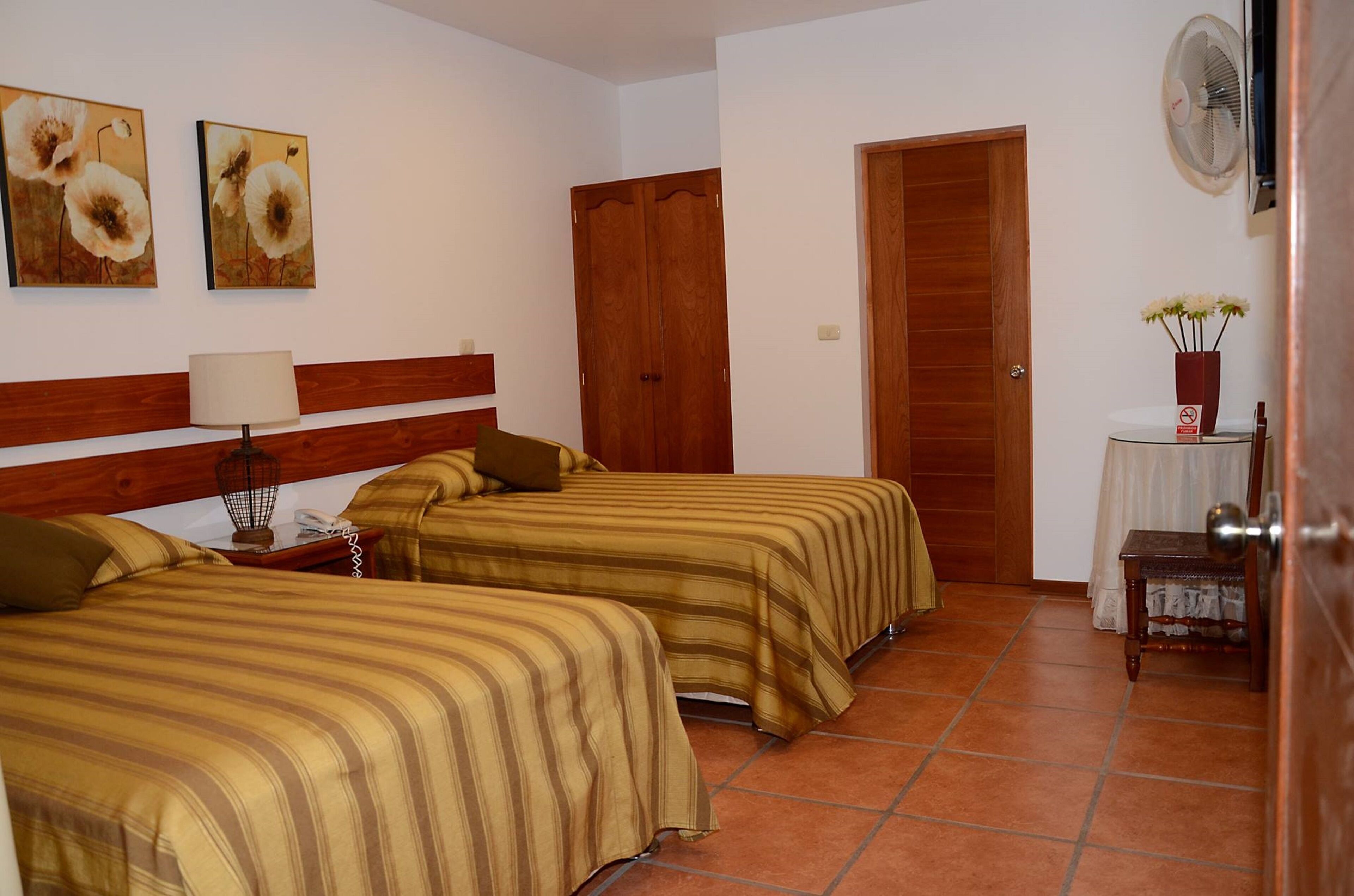 A hotel room with two beds, a wooden headboard, a nightstand with a lamp, a wooden door, a small table with a vase of flowers, and two wall-mounted paintings.