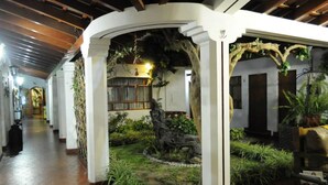A corridor with a covered outdoor area featuring a wooden structure, a small garden with plants and a decorative tree trunk, and a bench. - Hotel Oro Viejo (Nazca)