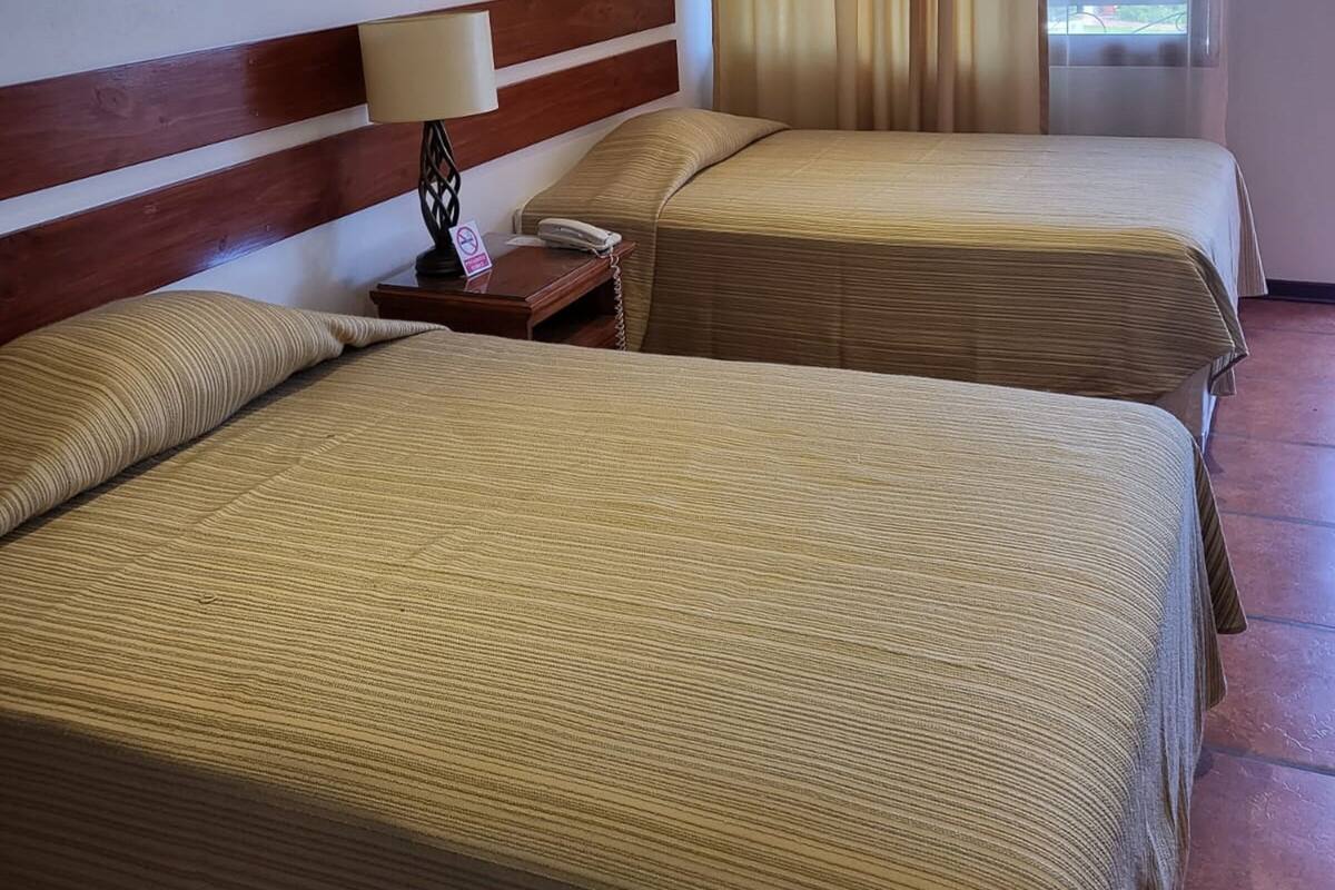 A hotel room with two beds, a wooden headboard, a nightstand with a lamp, and a window with curtains.