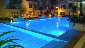 Piscina con sillas de descanso y palmeras, rodeada por un edificio de varios pisos con balcones.