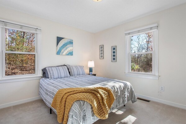 A bedroom with a bed, two windows, a nightstand, a lamp, and wall art.
