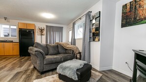 A living room with a grey sofa, a black ottoman, and a fluffy rug. A kitchen with wooden cabinets and a black refrigerator is visible in the background. - What a view!  (Victoria)