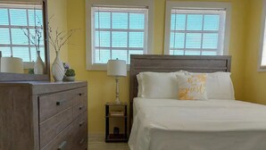 A bedroom with a wooden bed, a dresser, a nightstand with a lamp, and two windows with blinds.