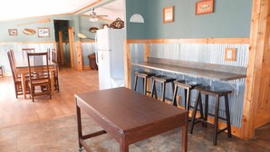 Interior rústico com uma mesa de jantar de madeira, banquetas de bar, uma geladeira e um ventilador de teto.