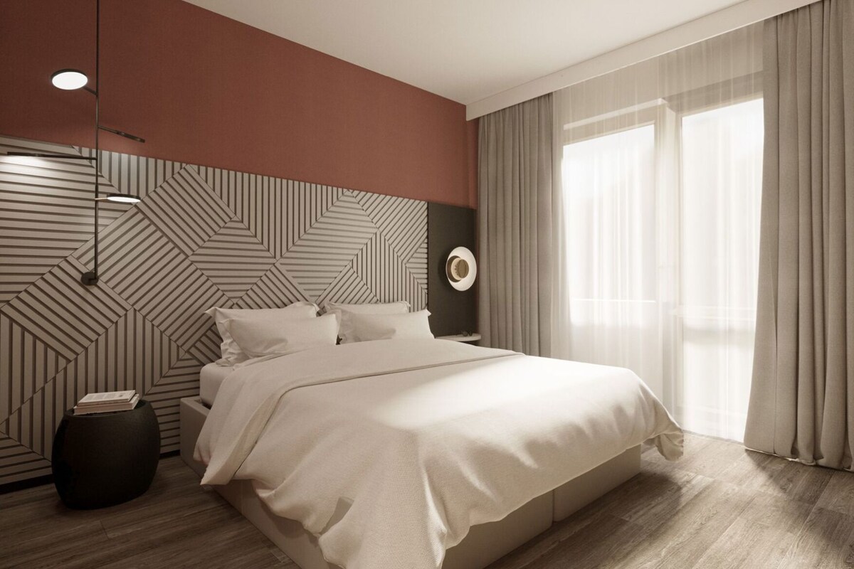 A modern bedroom with a geometric-patterned headboard, a large bed, a round side table, and a large window with curtains.
