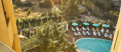 Un área de piscina en la azotea con sillones de descanso y sombrillas, rodeada de árboles y edificios.