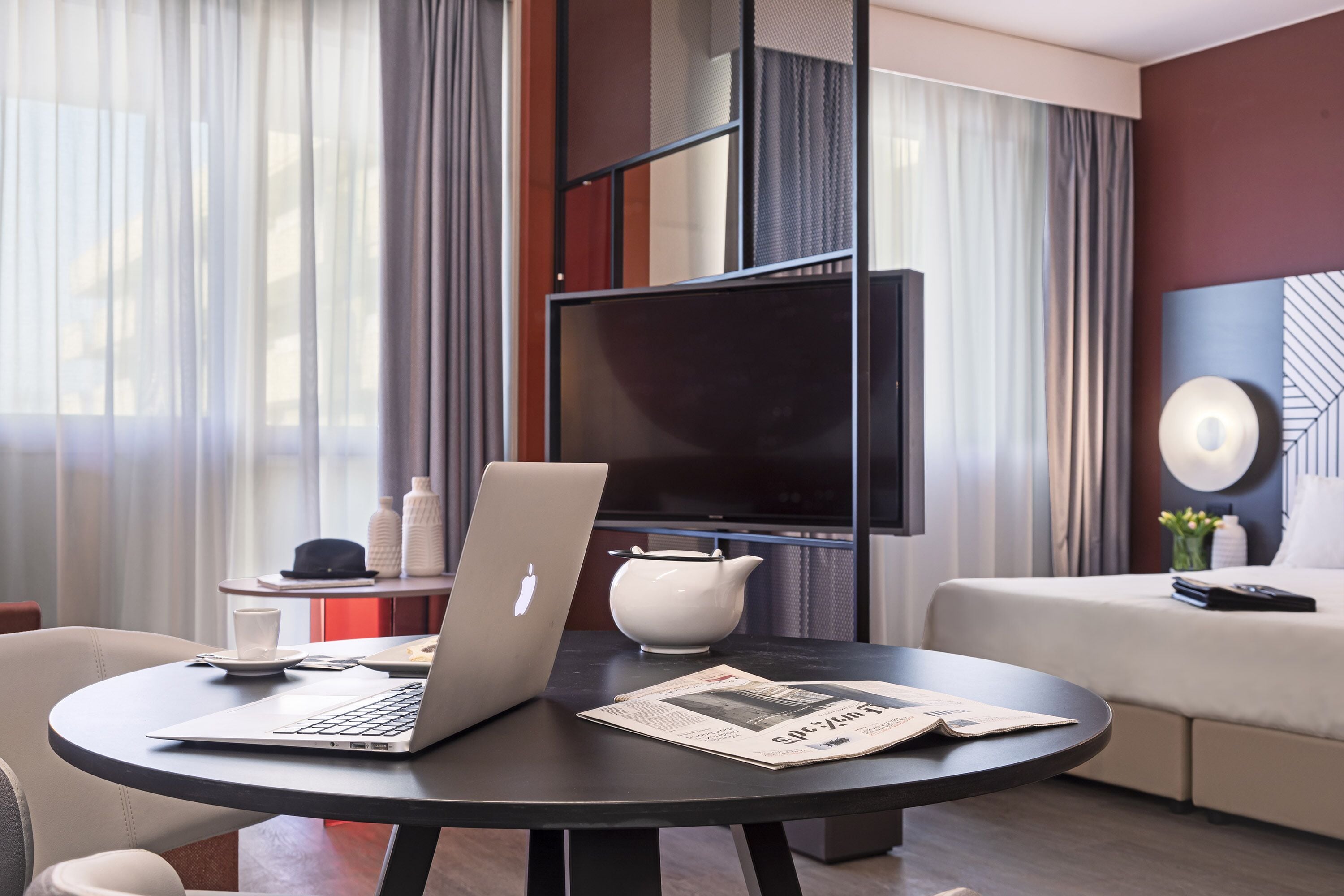 A modern hotel room with a round black table, a laptop, a newspaper, a television, and a sofa.