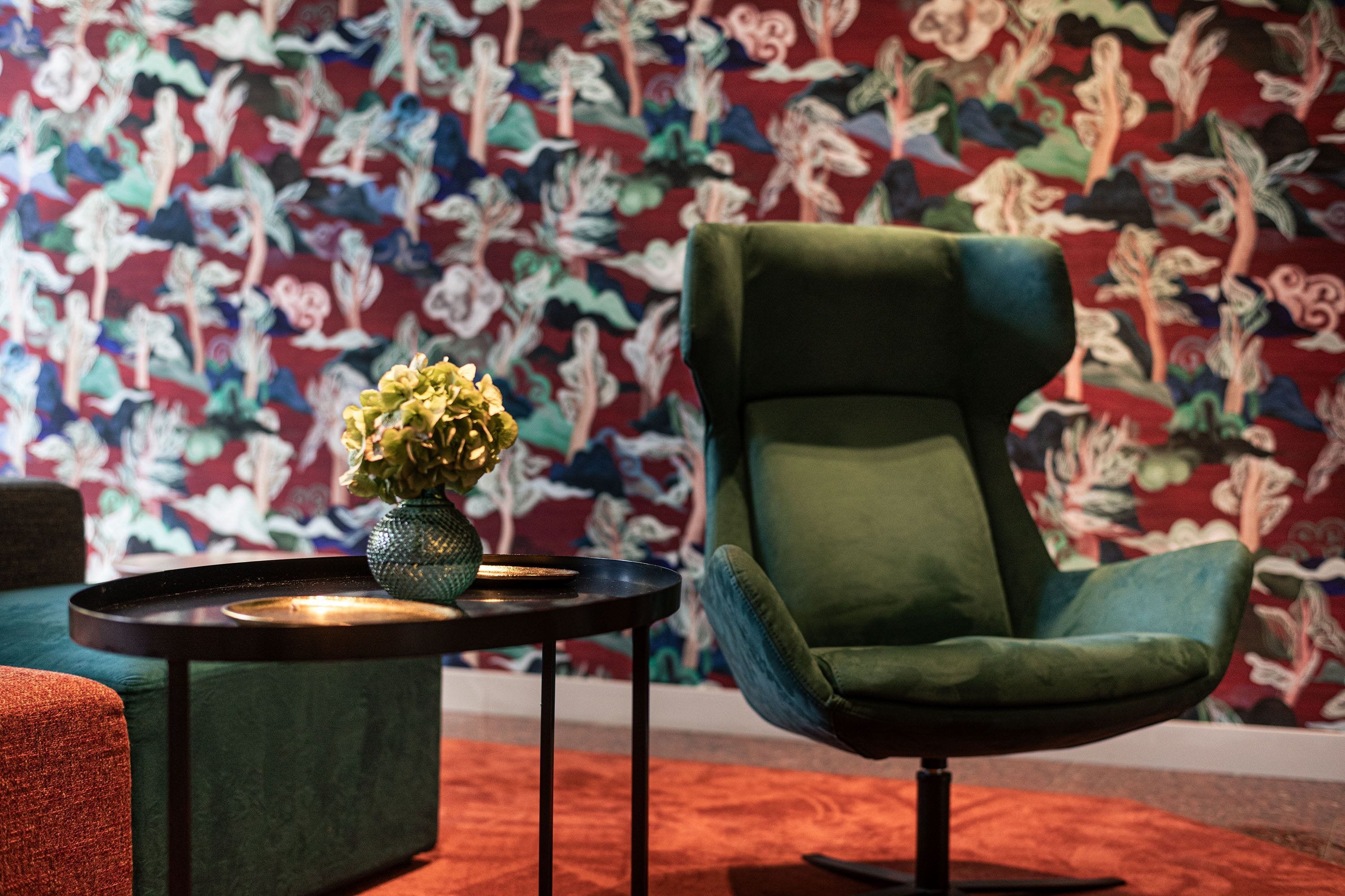 A room with a green armchair, a small table with a vase of flowers, and a patterned wallpaper.