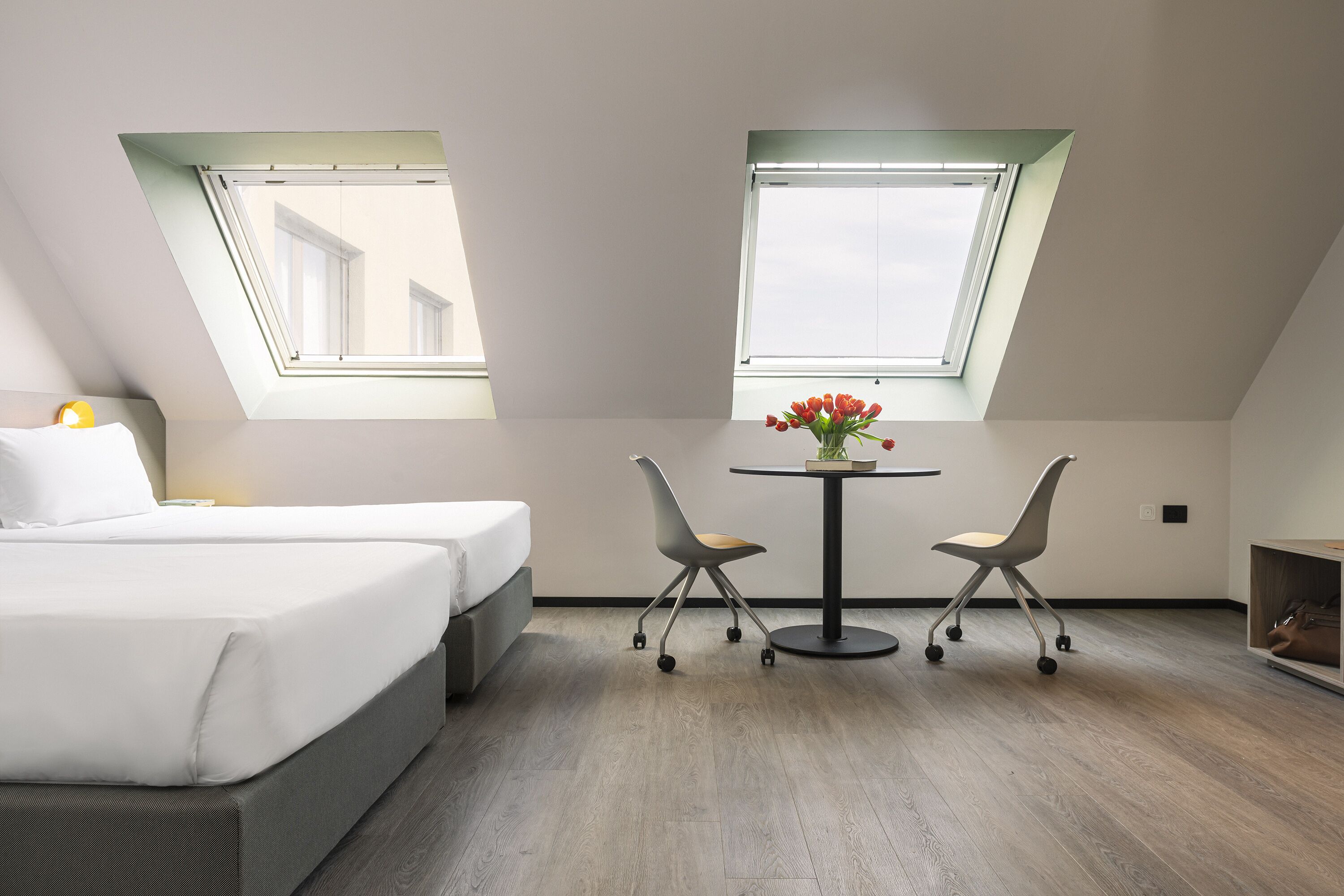 Quarto pequeno e moderno com uma cama, duas cadeiras, uma mesa e dois claraboias.