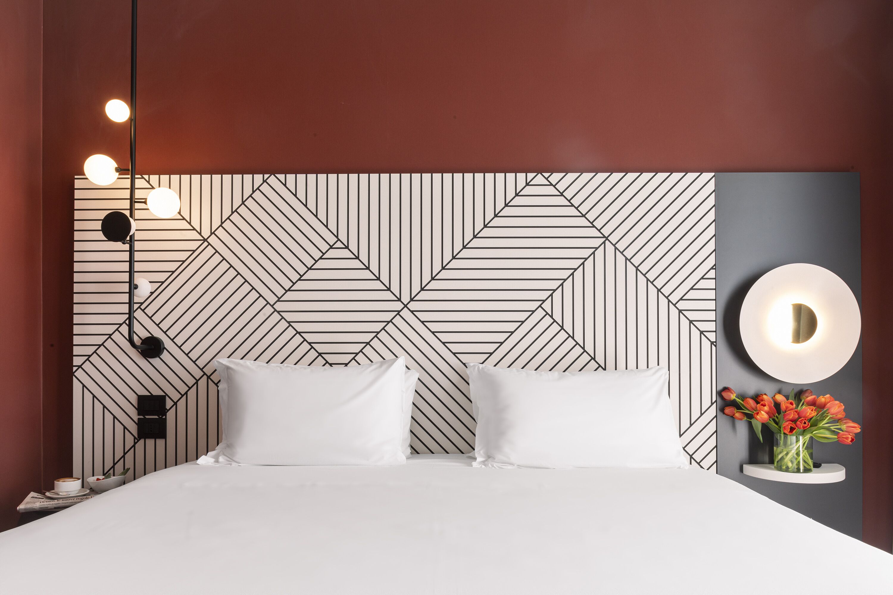 Une chambre d’hôtel moderne avec une tête de lit à motifs géométriques, du linge de lit blanc, une table de chevet avec une lampe et un vase rempli de fleurs rouges.