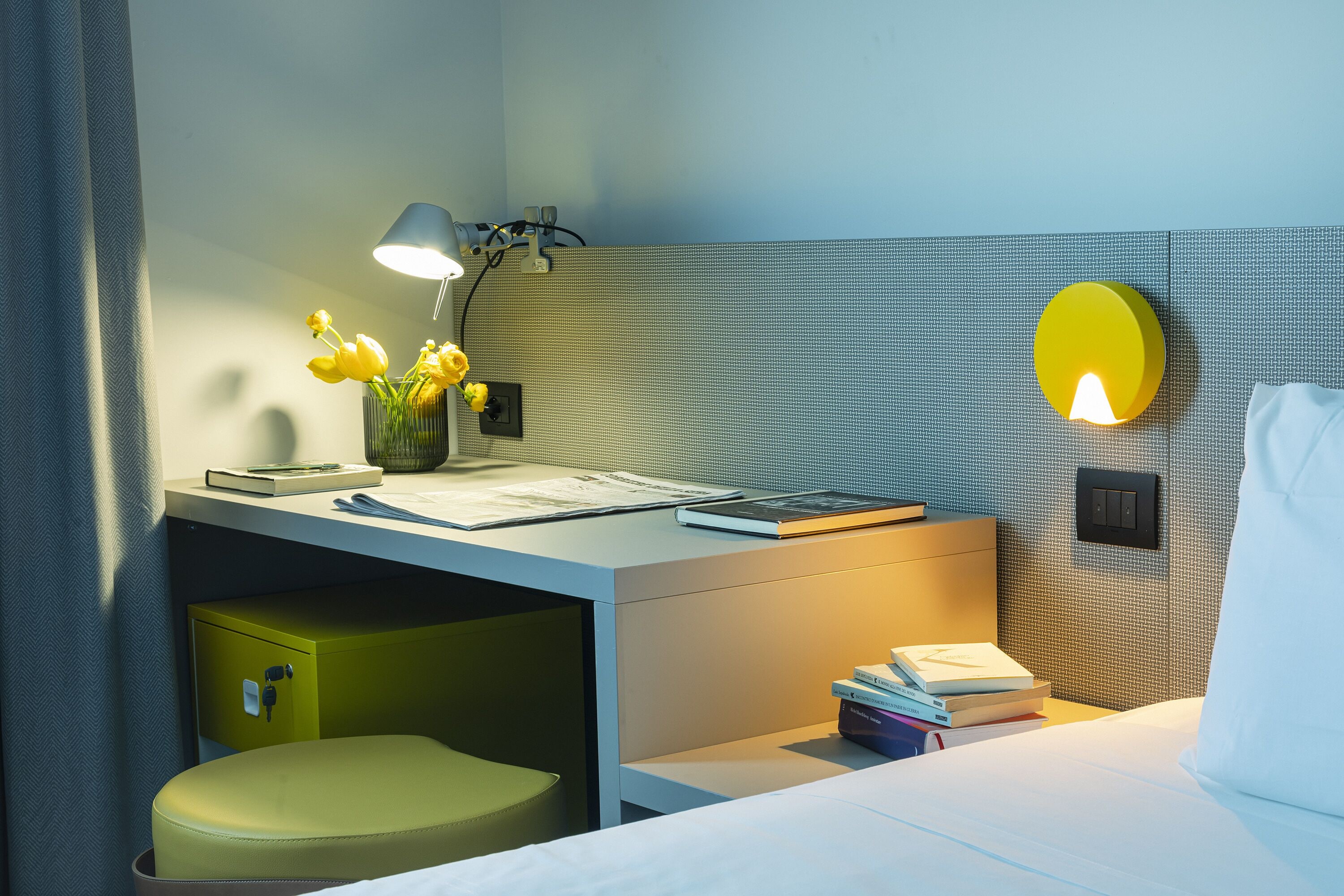 Une chambre d’hôtel moderne, équipée d’un bureau, d’une lampe, d’un vase avec des fleurs et d’un lit avec un oreiller.