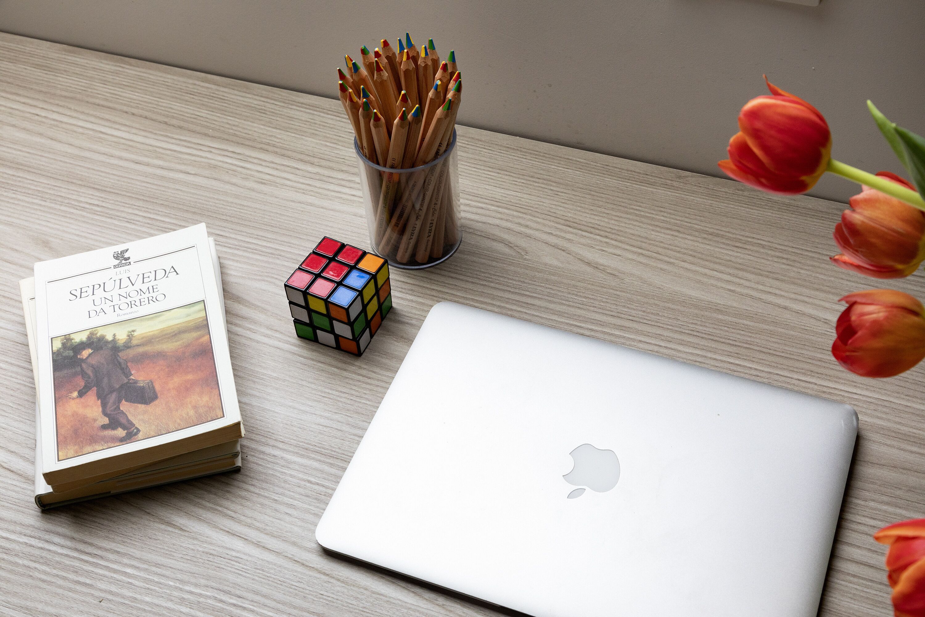 A laptop, a Rubik's cube, a book, a pencil holder, and tulips on a wooden surface.