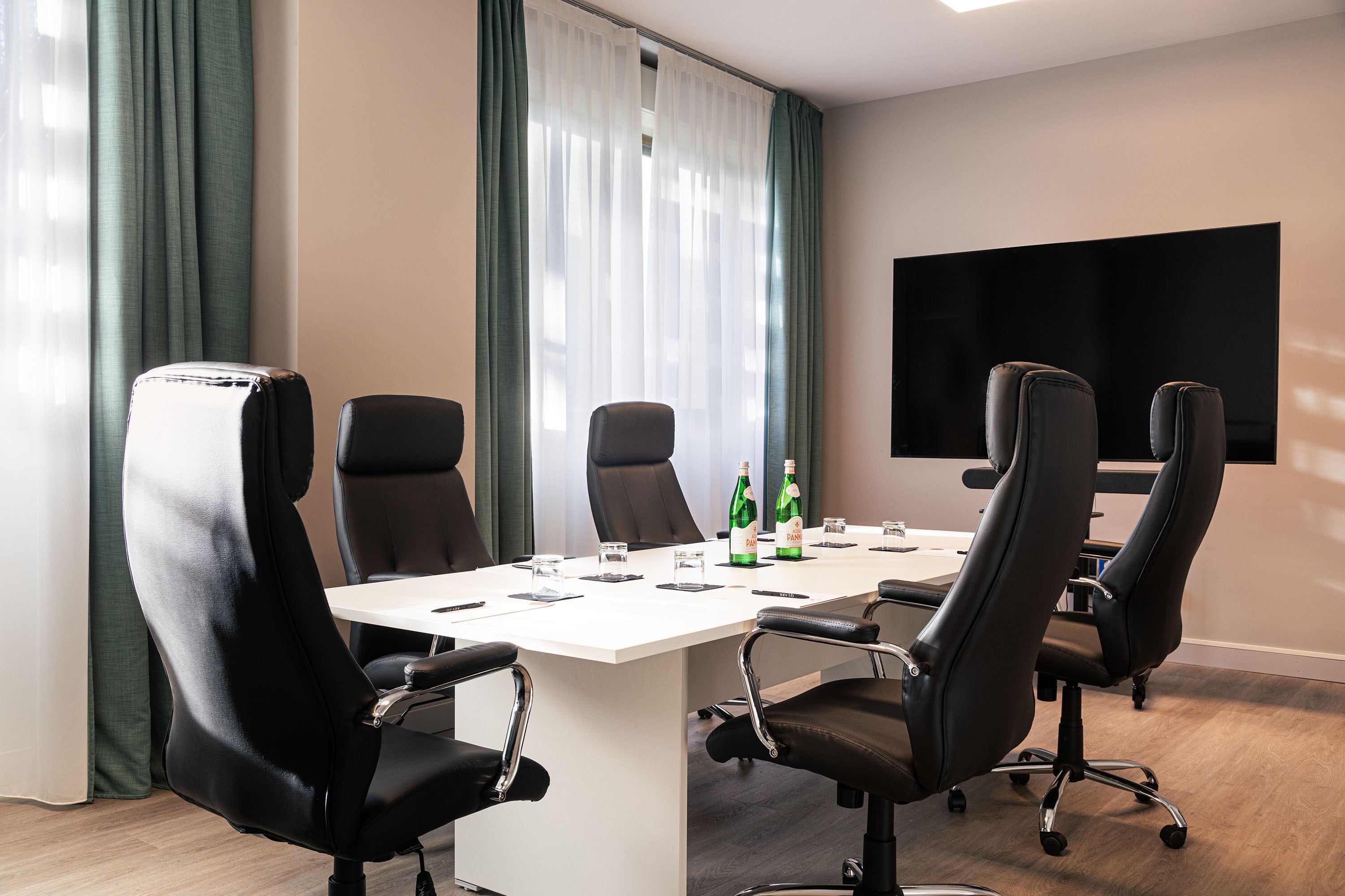 A conference room with a white table, black chairs, a flat-screen TV, and a window with green curtains.