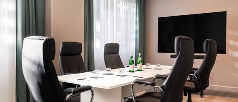 A conference room with a white table, black chairs, a flat-screen TV, and a window with green curtains.